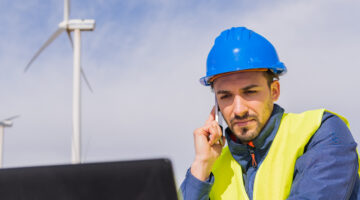 Worker With Safety Helmet And Reflective Vest, Talking On His Mobile Phone While Checking Data On His Laptop In An Electric Generator Park. Climate Change And Renewable Energy Concept.